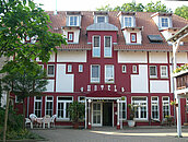 Blick auf den Eingang des Hotels. Die Außenfassade wurde in weiß und rot gestrichen. Über dem Vordach ist in weißer Schrift Hotel geschrieben.
