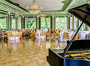 Ein lichtdurchfluteter Ballsaal der mit Tischen und einem Piano ausgestattet ist
