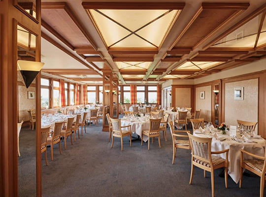 Weitläufiger Speisesaal mit vielen gemütlichen Tischgarnituren und hellen Fenstern, sowie schönen Deckenleuchten in hölzerner Ummantelung.