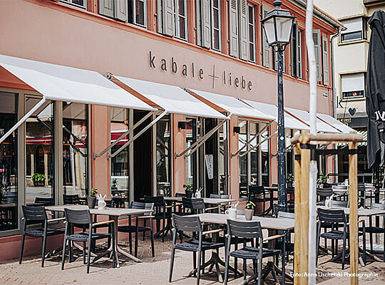 Außenansicht des k a b a l e  +  l i e b e mit Markise über Tischen an Sonnigen Tag