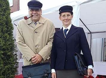 Zwei Schauspieler mit Aktentasche stehen nebeneinander und grinsen in die Kamera.