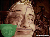 Ein mit Narben durchzogenes Gesicht hinter einem Glas mit giftig grüner Flüssigkeit 