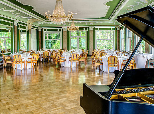 Ein lichtdurchfluteter Ballsaal der mit Tischen und einem Piano ausgestattet ist