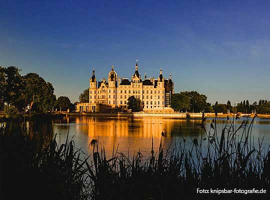 Krimidinner Schwerin | Krimidinner Schwerin im wunderschönen Schloss See mit Schloss im Hintergrund, Schloss wird von Sonnenlicht angestrahlt, strahlend blauer Himmel