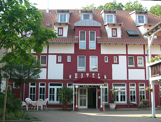 Blick auf den Eingang des Hotels. Die Außenfassade wurde in weiß und rot gestrichen. Über dem Vordach ist in weißer Schrift Hotel geschrieben.