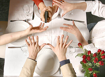 Vier Personen strecken Ihre Hände über einem gedeckten Tisch aus, eine Person hält eine Waffe beim spannenden Impro Krimidinner