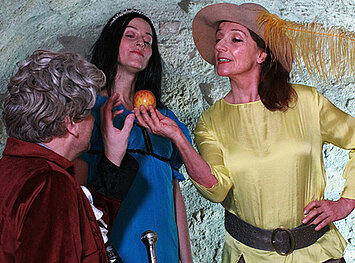 Zwei Frauen und ein Mann stehen zueinander gekehrt. Eine Frau mit Hut hält einen Apfel in ihrer Hand, dieser wird von allen drei Personen begutachtet. 