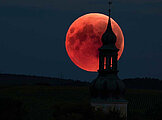 Leuchtend roter Mond am Nachthimmel, etwas verdeckt durch einen Kirchturm