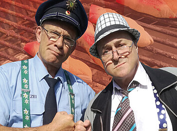 Ein Polizist und ein Mann mit gemusterten Hosenträgern, Hut und Krawatte geben sich die Faust. 