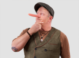 Michael Eller, der Comedian, in brauner Weste und Schirmmütze. Er hat eine lange Pinocchio Nase umgeschnallt.