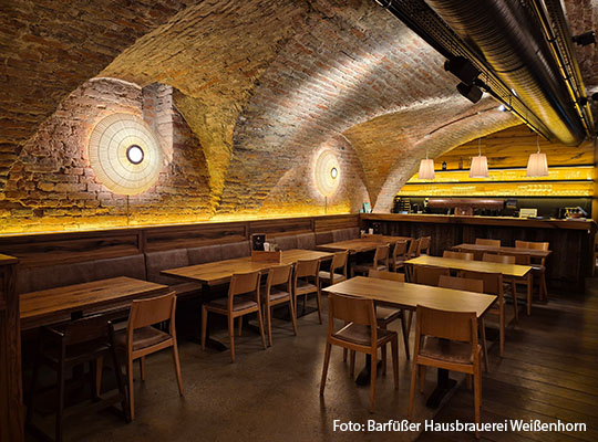 Langer Raum mit Lampen an der Wand, mit Holztischen und Stühlen im inneren. Hinter den Tischen ist eine Bar.