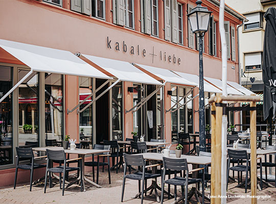 Außenansicht des k a b a l e  +  l i e b e mit Markise über Tischen an Sonnigen Tag