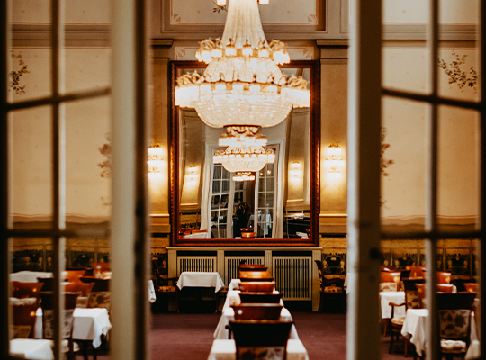 Durch Fenster fotografierter Saal mit Tischen und Stühlen sowie imposantem Kronenleuchter