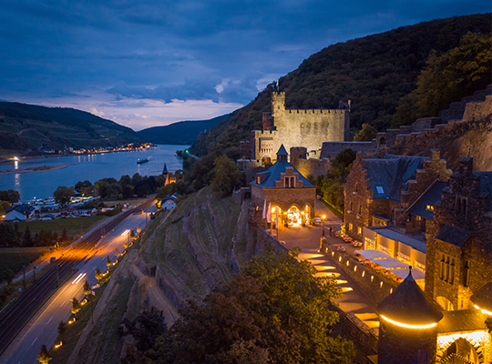 Seitenansicht bei Nacht auf die Burg Reichenstein mit dem Rhein daneben. Bei Dunkelheit ist die Burg hell beleuchtet.