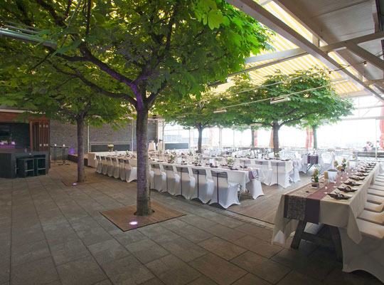 Große Terrasse mit schönen Baumen und riesiger Jalousie und feierlich angerichteten Esstischen