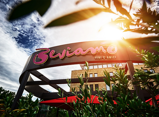 Außenansicht des Restaurants mit Blick auf das schön designte Eingangsschild mit der Aufschrift "Beviamo VINI E AMICI". Im Hintergrund ist ein heller Sonnenschein mit blau bewölktem Himmel zu sehen. 