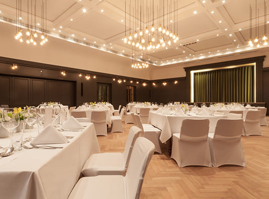 Großer Speisesaal mit wundervollen Deckenleuchten und mit weißen Hussen dekorierten Tischen. Zudem ein riesiger Bühnenbereich mit grünem Vorhang.