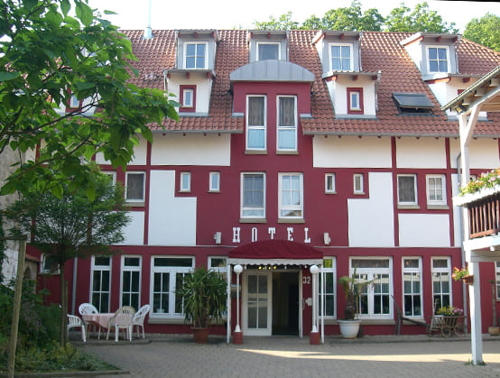 Blick auf den Eingang des Hotels. Die Außenfassade wurde in weiß und rot gestrichen. Über dem Vordach ist in weißer Schrift Hotel geschrieben.