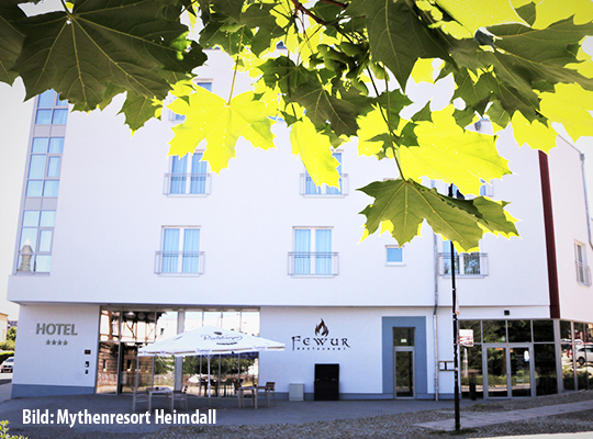 Außenansicht des Hotels und Krimidinner Restaurants inklusive Terrasse und grünen, in das Bild hängenden Blättern 