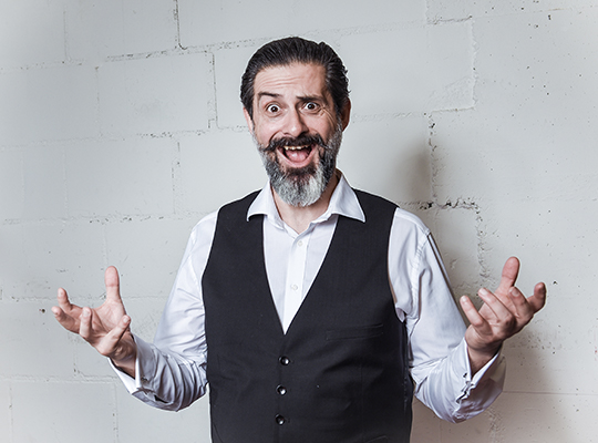 Der Schauspieler steht vor einer grauen Wand, hält seine Hände nach oben geöffnet und zieht dabei eine lustige Grimasse. 