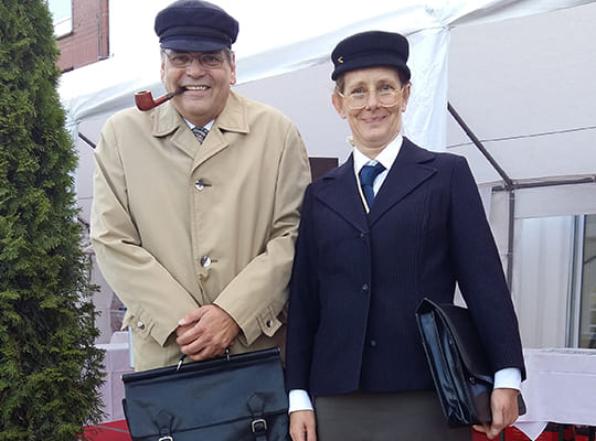 Zwei Schauspieler mit Aktentasche stehen nebeneinander und grinsen in die Kamera.