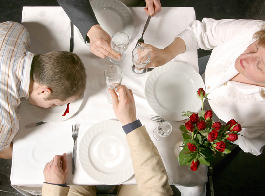 Spannung beim Krimidinner: Drei Personen stoßen über einem gedeckten Tisch an, eine weitere Person liegt mit dem Kopf auf dem Teller auf dem eine Blutspur zu sehen ist 